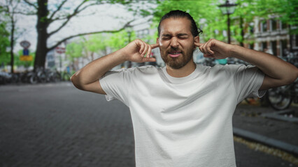 Naklejka premium Man with beard pressing fingers into ears on a crowded city street, eyes closed and grimacing; frustration overload.