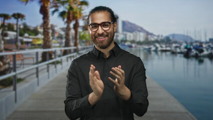Man clapping hands on a street pier at a marina, wearing glasses and beard and smiling into camera;...