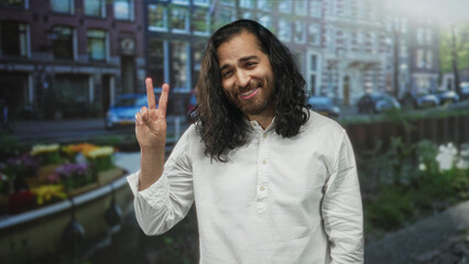 Fototapeta premium Man smiling and holding up two fingers in a peace sign on amsterdam street by a canal and buildings; cheerful.