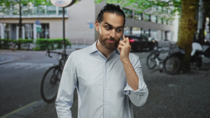Man with beard and rolled sleeves holding smartphone to ear on a busy city street near parked bicycles and cars; thoughtful business call.