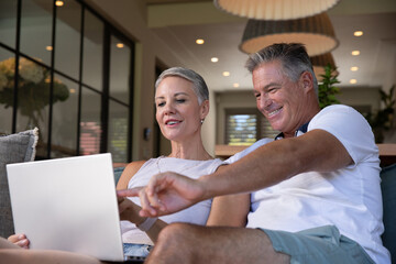 Senior couple sitting on couch in open-plan room using silver laptop, pointing at screen