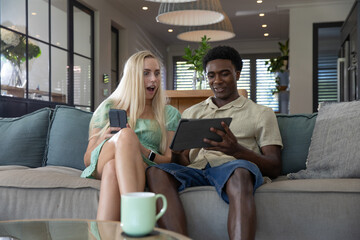 Diverse couple sitting on couch reacting to smartphone while man holding tablet in living room