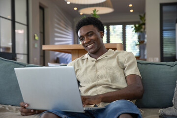 African american man working on laptop on teal sofa in living room with pendant lights
