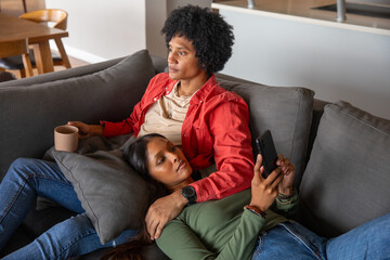 Diverse couple wearing smartwatch holding mug and smartphone while relaxing on couch in living room