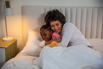 Mother and daughter lying together in bedroom on bed hugging under white duvet next to bedside lamp