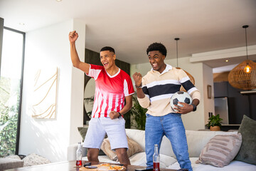 Diverse male friends wearing soccer jersey cheering at home by coffee table with pizza, soccer ball