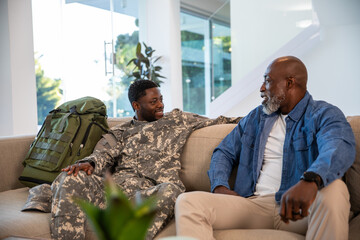 African american father and son sitting on sofa at home in military uniform with backpack