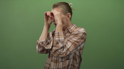 Woman using hand binoculars with exposed forearms, puckered lips and plaid shirt in green studio;...