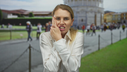 Fototapeta premium Young blonde woman experiencing tooth pain near pisa tower outdoors in italy, displaying a grimace, dressed in a white shirt, with a scenic background of tourists and historical architecture.