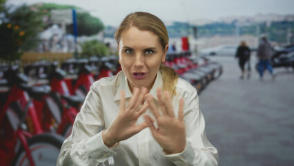 Woman gesturing energetically in a white shirt at an outdoor bicycle parking area with blurred bicycles and trees in the background, capturing a lively moment.