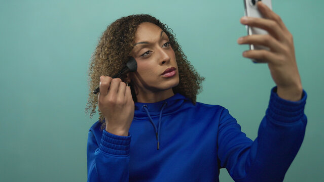 Woman brushes cheek with makeup brush while holding smartphone in studio; confidence selfcare empowerment identity. - Powered by Adobe