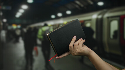 Caucasian man holding a book at a bustling indoor train station with blurred passengers and trains in the background.