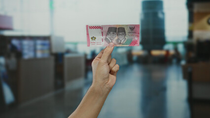 Hand holding indonesian rupiah at airport terminal illustrating travel and finance with focus on banknote indoors by a man showcasing currency in busy surroundings.