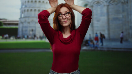 Redhead woman in red sweater playfully poses with hands as ears in front of pisa tower outdoors on a sunny day in pisa, capturing a fun travel moment.