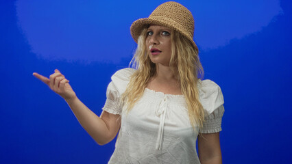 Woman points finger while wearing straw hat and white shirt in blue studio with vivid backdrop;...