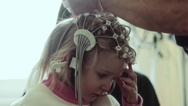 Close up view of adult hands adjusting electroencephalography cap on head of 4 years old Caucasian child. Child remains still, expression of concentration during neurological examination.