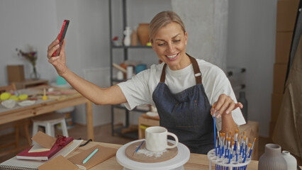 Woman holding smartphone with outstretched arm, gesturing to a ceramic mug on a potter's wheel in a...