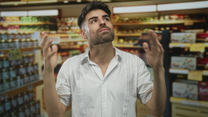 Fototapeta premium Man with raised hands and pursed lips wears white shirt at grocery shelves in a supermarket aisle; frustration irritation stress annoyance.
