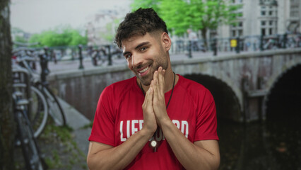 Man holds hands pressed together while smiling in a red lifeguard shirt on a street beside a canal bridge; dedication.