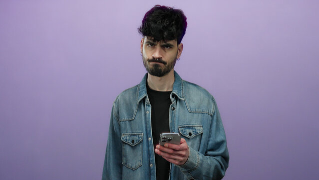 Young man with smartphone looking frustrated against isolated purple background expressing modern digital frustration with a stylish appearance and a contemplative pose.