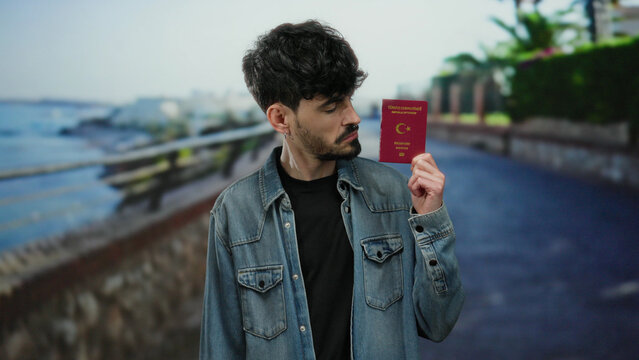 Young man holding turkish passport on seaside promenade with ocean view and outdoor setting, conveying travel and identity themes in a relaxed outdoor environment.