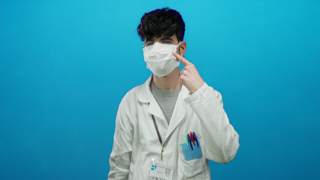 Young man in a white coat wearing a mask points at his face against a blue background, emphasizing medical awareness and responsibility.