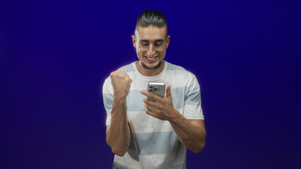 Man holding smartphone clenches fist and smiles while looking at the screen in a studio purple backdrop; triumph celebration.