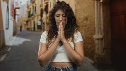 Naklejka premium Woman pressing hands together in prayer with closed eyes on a narrow old town street; solemn prayer.