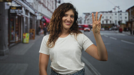 Woman holds up three fingers with smiling face wearing white tshirt and denim jeans on a busy street in the city; happiness confidence.