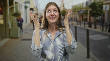 Fototapeta premium Woman with clasped hands smiling on street wearing striped shirt by storefronts and lampposts on sidewalk under city buildings in daylight; happiness.