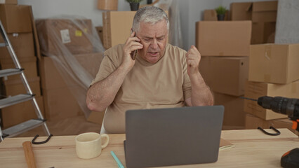 Man holds phone to ear and gestures at laptop amid packed moving boxes and tools in a building; moving frustration.