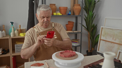 Man potter taps a red smartphone with his hand while seated at a pottery wheel shaping clay in a ceramics studio; craft focus patience.
