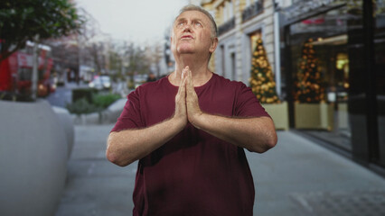 Senior man with hands pressed in prayer at chest on a busy city street sidewalk beside a storefront and christmas tree, looking upward in maroon shirt; faith hope.