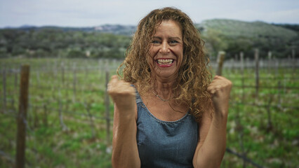 Obraz premium Woman clenches fists and smiles in denim dress amid a vineyard with wooden trellis in forest hills; joy triumph.
