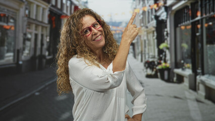Woman pointing finger upward and smiling with red glasses and white blouse on a street; joy confidence.