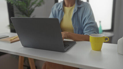 Woman at laptop clasping hands near chin wearing glasses and a denim shirt in a building; contemplation calm focus.
