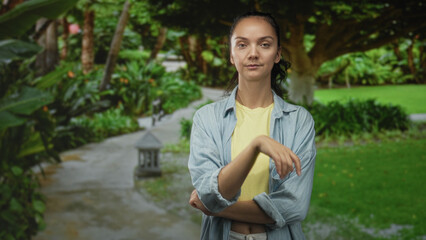 Fototapeta premium Woman standing with arms crossed showing bare forearms, wearing yellow shirt and denim overshirt on a garden path amid lush trees in forest; calm reflection.