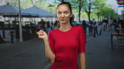 Woman in red shirt raises index finger in a lively street with outdoor cafe tents and passing...