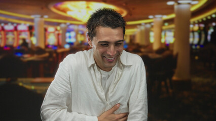 Fototapeta premium Smiling young hispanic man in white shirt enjoys his time inside a vibrant indoor casino filled with bright slot machines and lively atmosphere.