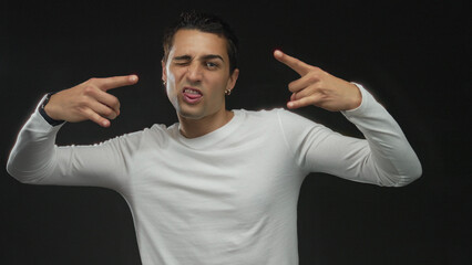 Naklejka premium Young hispanic man in white shirt shows rock horns hands gesture in studio; playful metal music spirit.