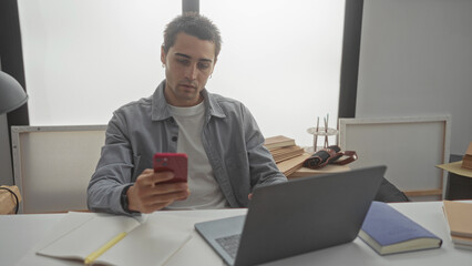 Young hispanic man sitting in a living room, engaged with a smartphone and laptop, surrounded by books and papers, creating a relaxed work-from-home atmosphere.