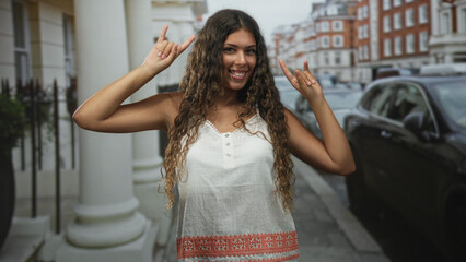 Naklejka premium Hispanic woman flashing rock horns with both hands on a residential city street by parked cars; playful joy.