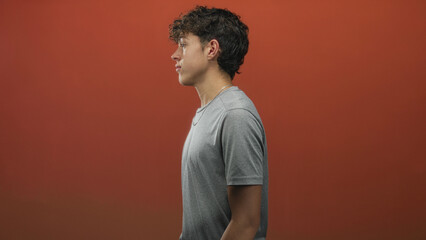 Teenage boy standing in profile with neutral expression, wearing grey t shirt and silver necklace, exposed neck visible in studio; calm introspection.