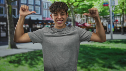 Teenage boy in gray tshirt points thumbs to chest on a busy city street plaza, smiling with raised...