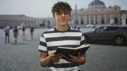 Teenage boy with glasses holds an open book and reads in front of a historic basilica building in a...