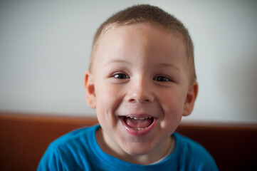 Young boy smiles widely. A boy with short light hair sits on a wooden bench. He has a big smile on his face that shows happiness. The room has natural good lighting coming from a window