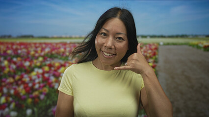 Fototapeta premium Woman smiling and showing call gesture with hand and ring while wearing yellow t shirt on a studio tulip field set; friendly joy.