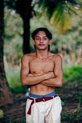 Portrait of a teen in traditional attire with arms crossed, standing in a palm grove with soft natural light.