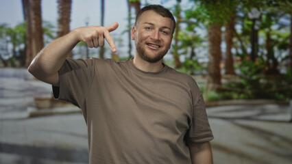 Man points finger down with bare forearm visible on street wearing brown t shirt and smiling at camera in outdoor plaza; friendly invitation.