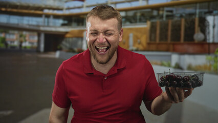 Man holding plastic container of cherries laughing with eyes closed in building courtyard; joy simple pleasure.